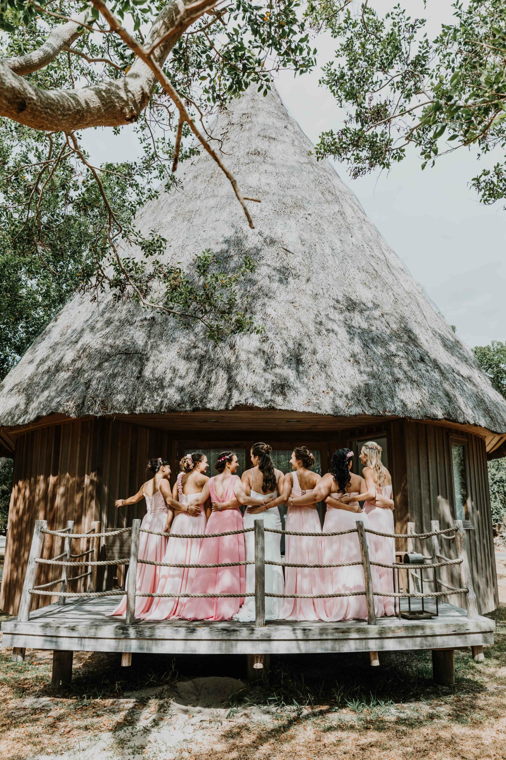 la mariée et les demoiselles d'honneur sont en ligne, de dos, et se regardent sur la terrasse d'un bungalow en bois en bord de plage