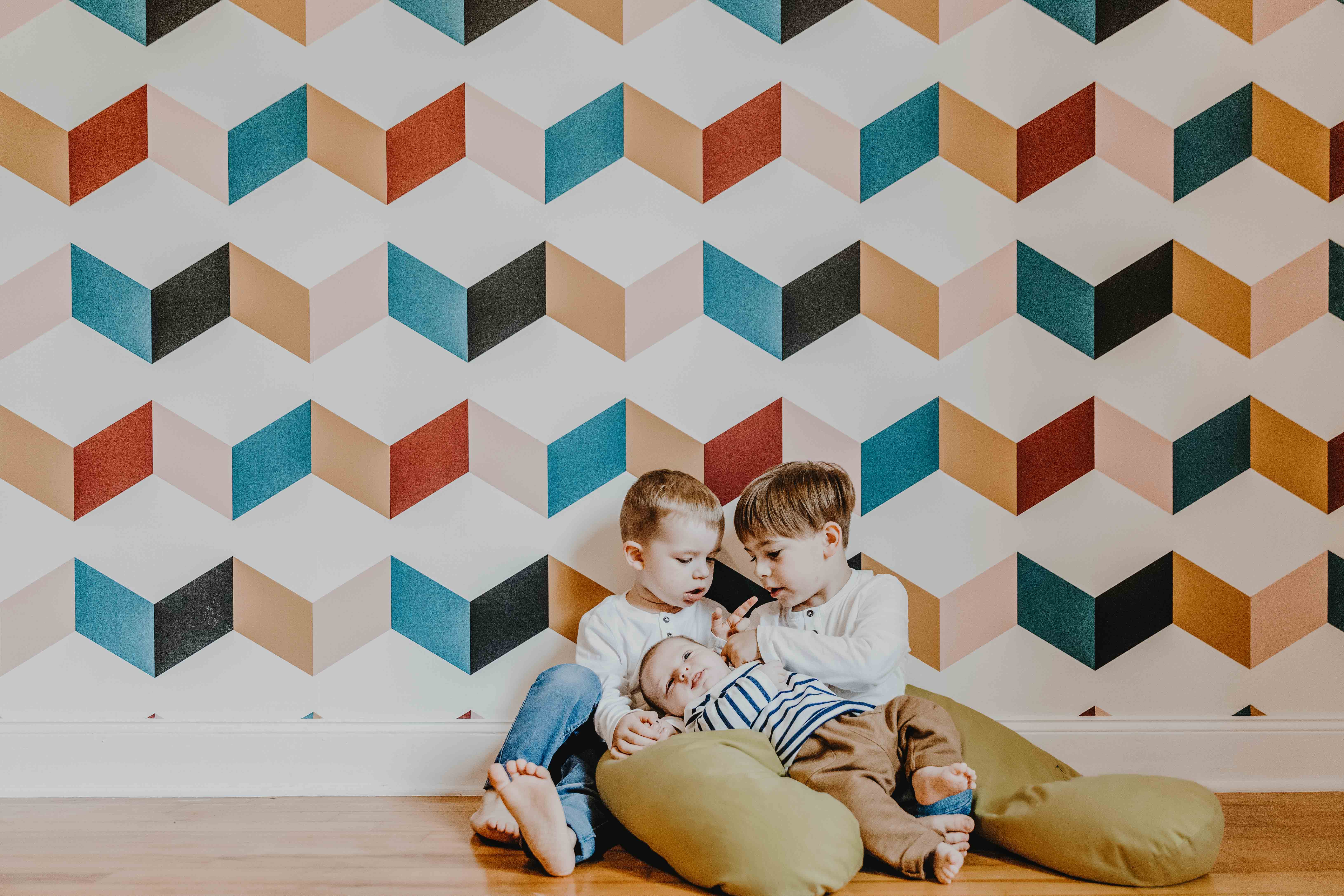 séance photo famille lifestyle à domicile. les enfants sont assis par terre contre un mur à tapisserie graphique et colorée. les grands frère tiennent bébé dans leurs bras et le regardent
