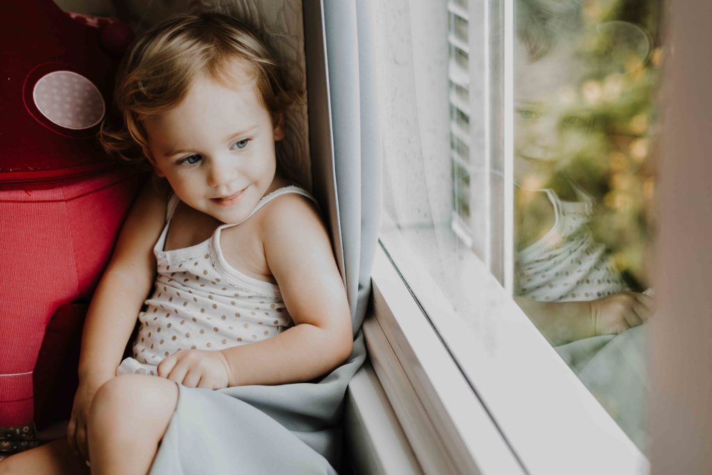 photographe famille lifestyle à domicile, immortaliser des moments de complicité et de douceur dans votre environnement quotidien. L'enfant regarde par la fenêtre de sa chambre à Colmar.