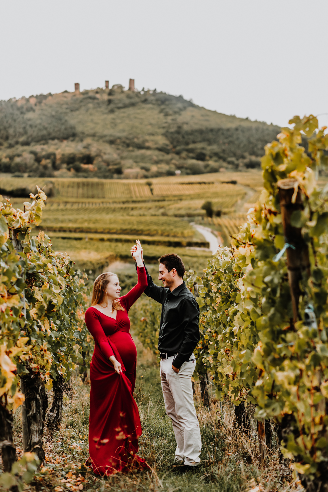 une femme enceinte dans une robe rouge danse avec son mari dans les vignes en automne