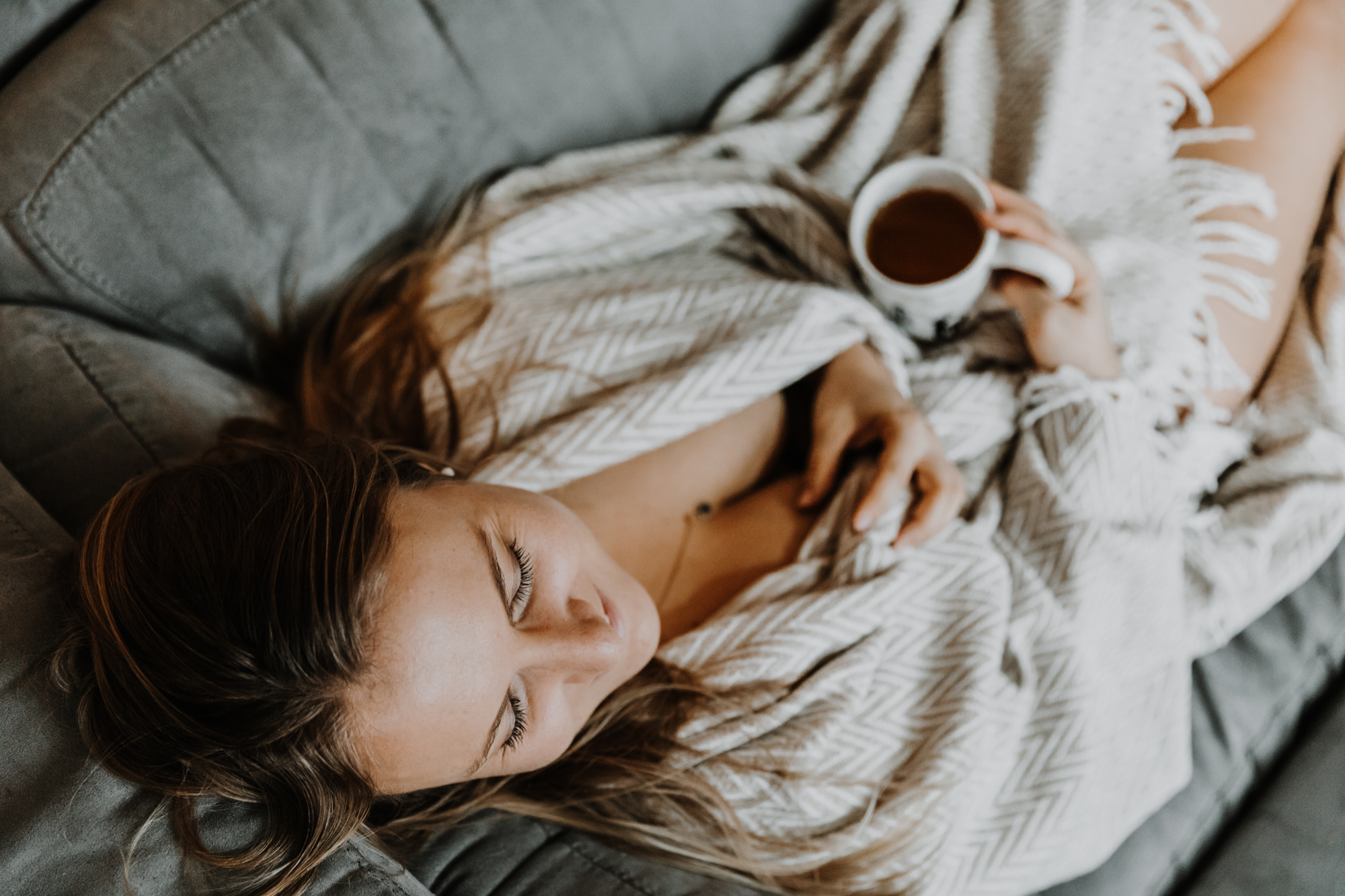 jeune femme allongée sur son canapé dénudée sous sa couverture, tient une tasse de thé et passe la main dans ses cheveux