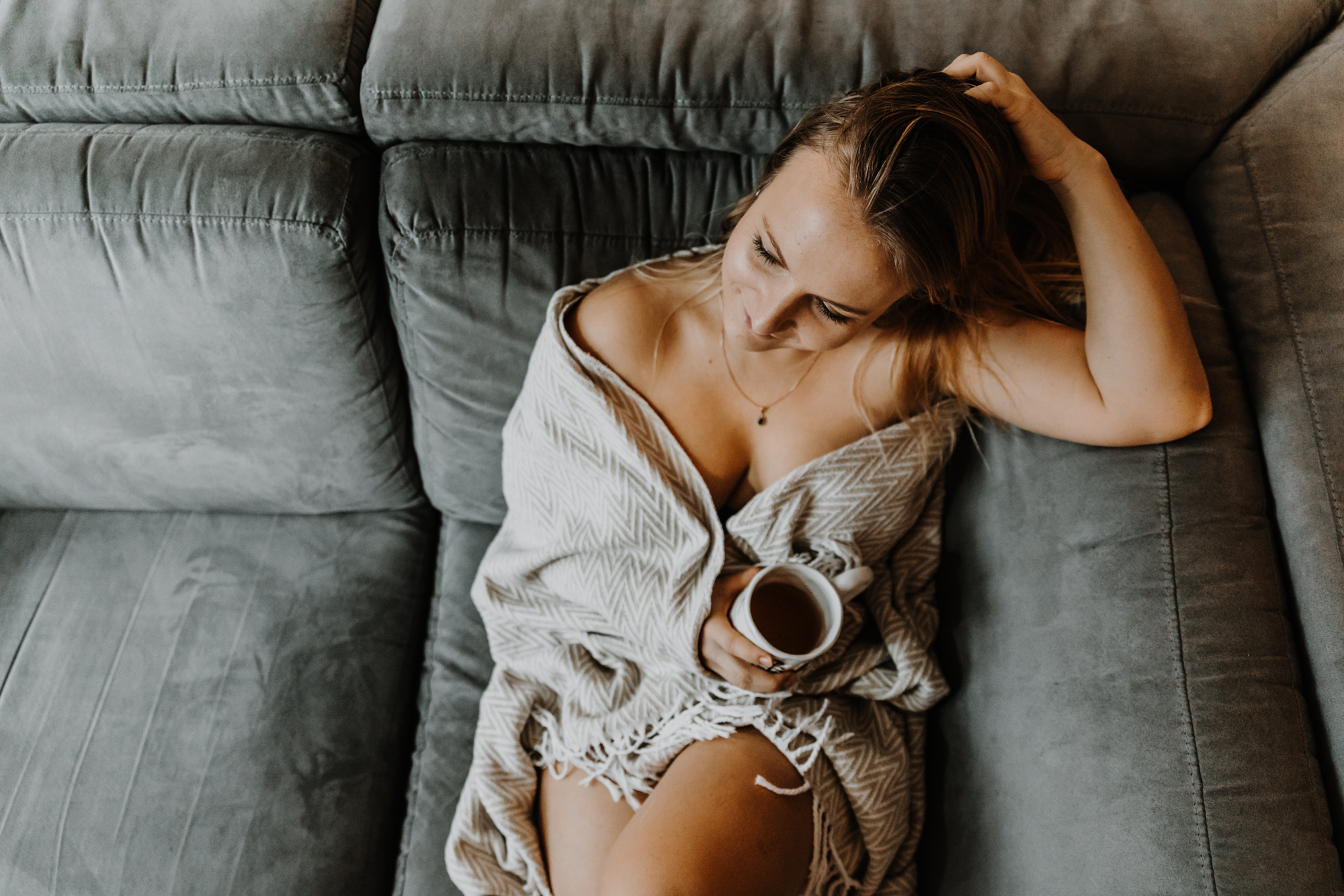 jeune femme allongée sur son canapé dénudée sous sa couverture, tient une tasse de thé et passe la main dans ses cheveux