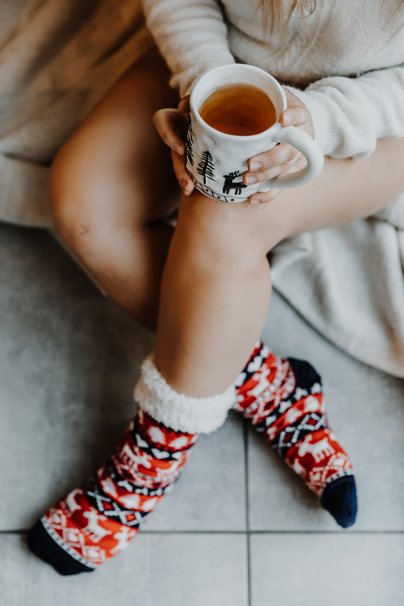 détail des jambes d'une jeune femme assise sur une couverture au sol, les jambes nues dans des chaussettes de noël, tenant une tasse de thé