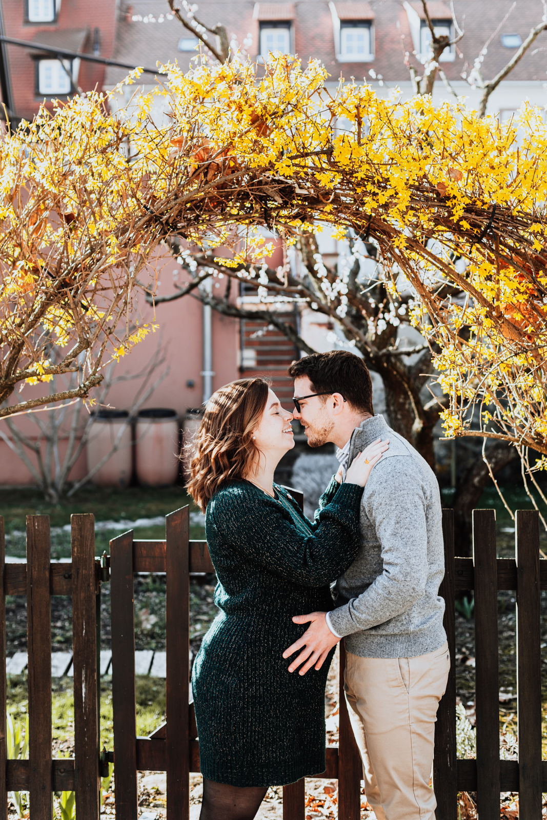 un couple se tient debout sous une arche de fleurs jaune, l'homme et derrière la femme et a les mains posées sur le ventre se sa femme enceinte