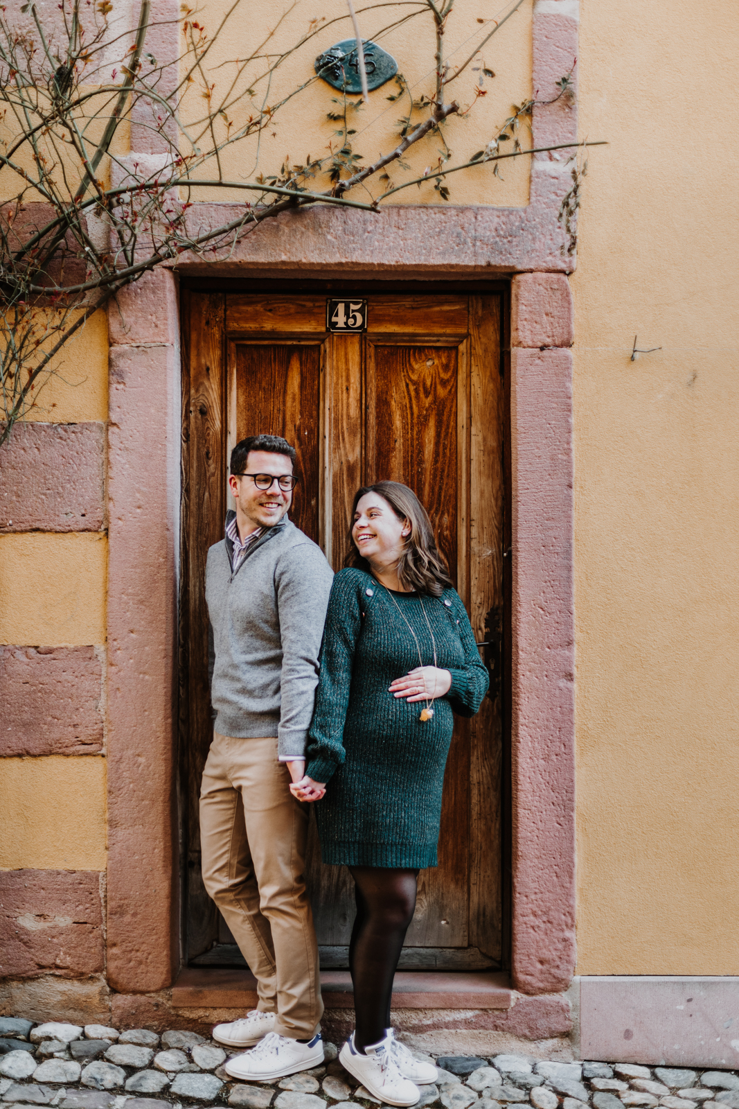 une homme et une femme enceinte, dos à dos devant une ancienne porte en bois dans le village de Kaysersberg en alsace