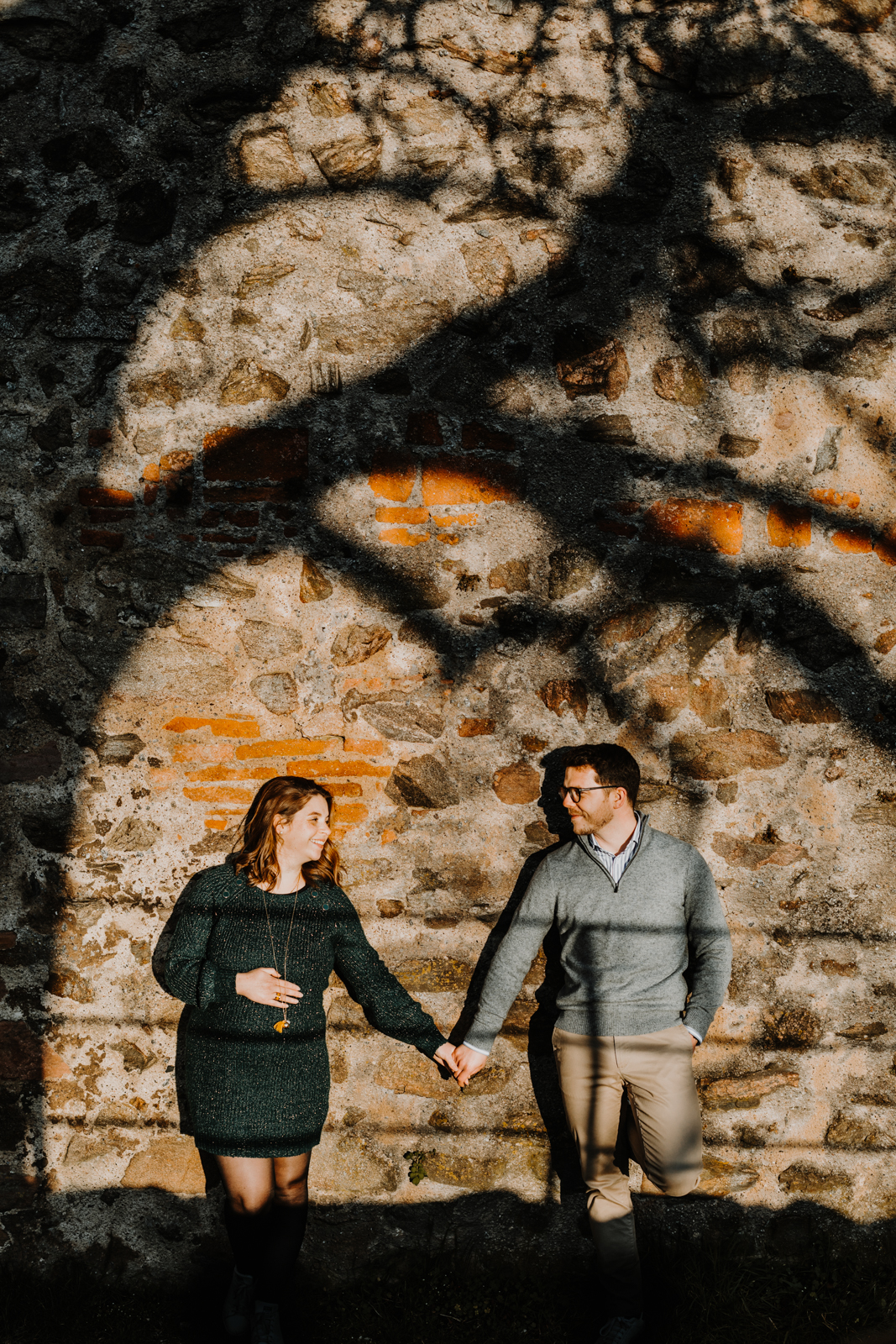couple se tenant par la main, adossés contre un mur en pierres, l'ombre d'un arbre projetée sur le mur