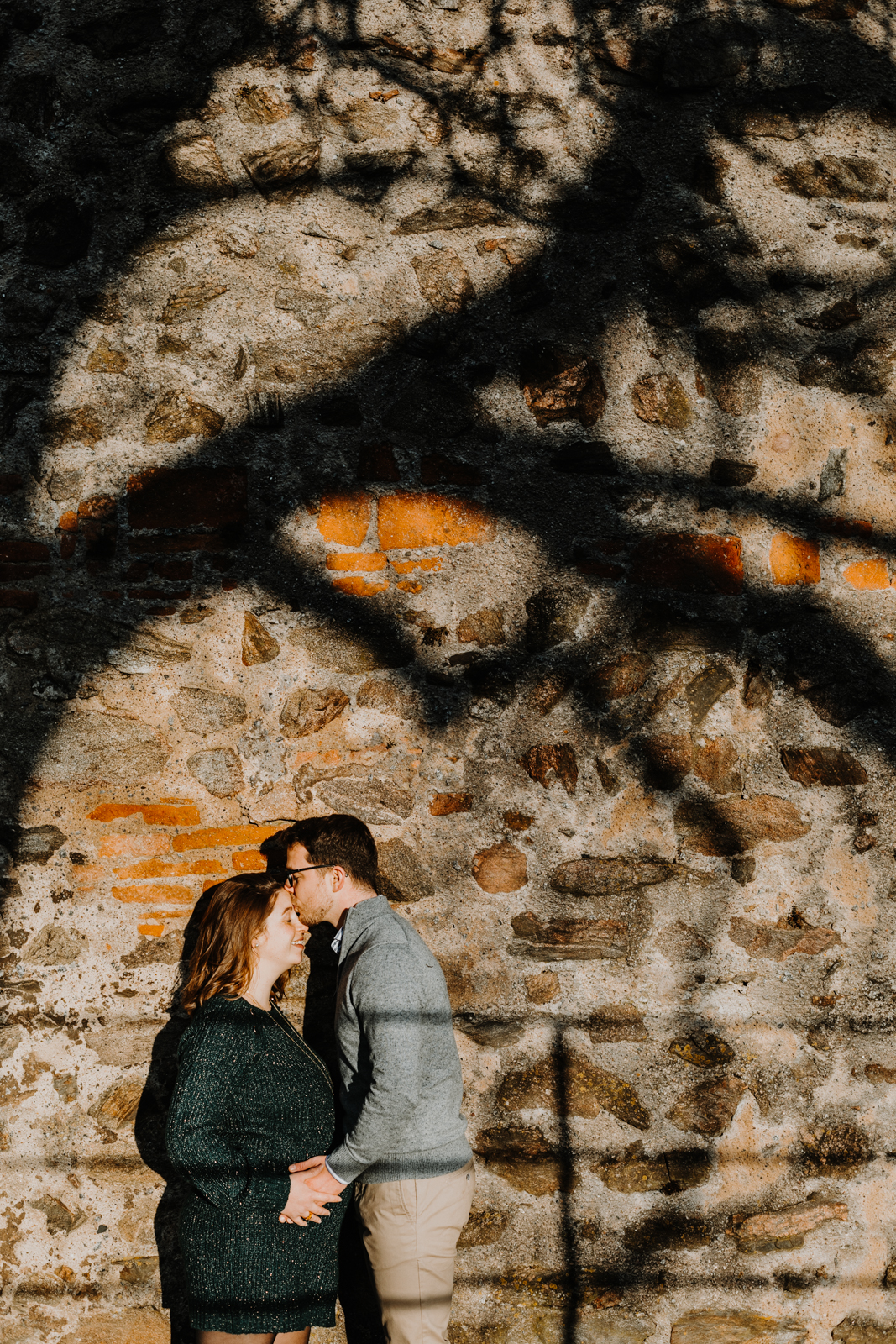 couple de profil contre un mur en pierres, l'homme embrasse la femme enceinte sur le front, l'ombre d'un arbre est projetée sur le mur