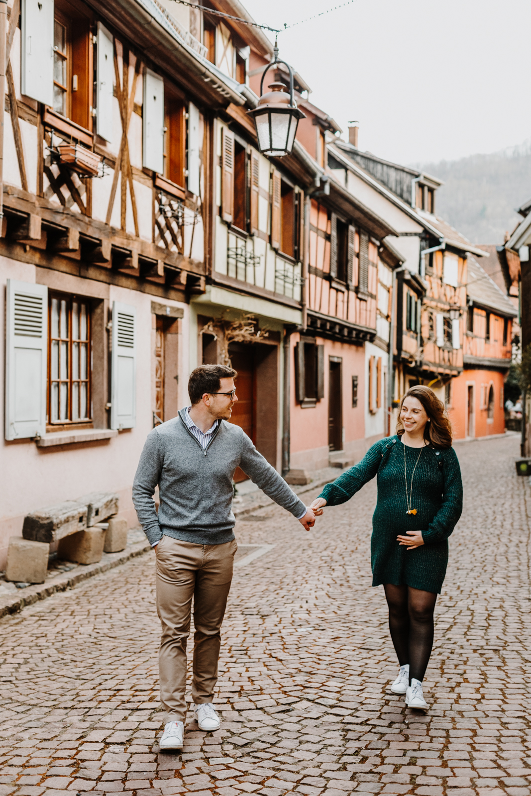 un homme et une femme enceinte marchent main dans la main dans une ruelle pavée du village de Kaysersberg en alsace