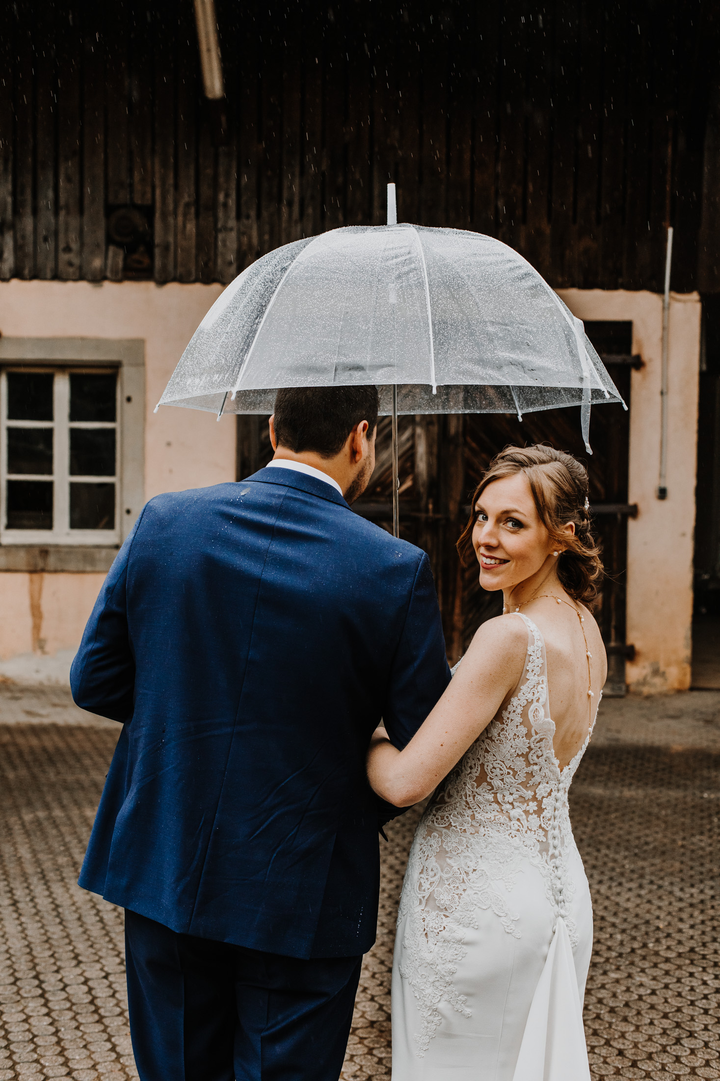 les mariés marchent sous la pluie, dans une ancienne cour, sous un parapluie transparent