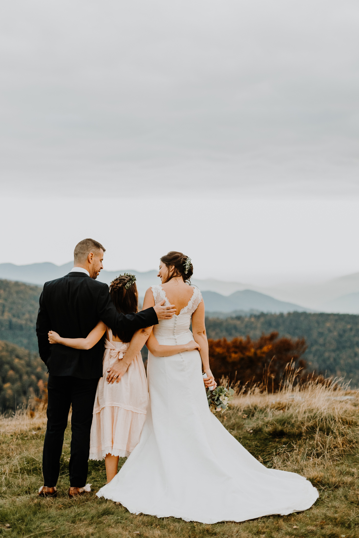 le couple de mariés et leur fille de dos, bras dessus bras dessous face à un paysage de montagne