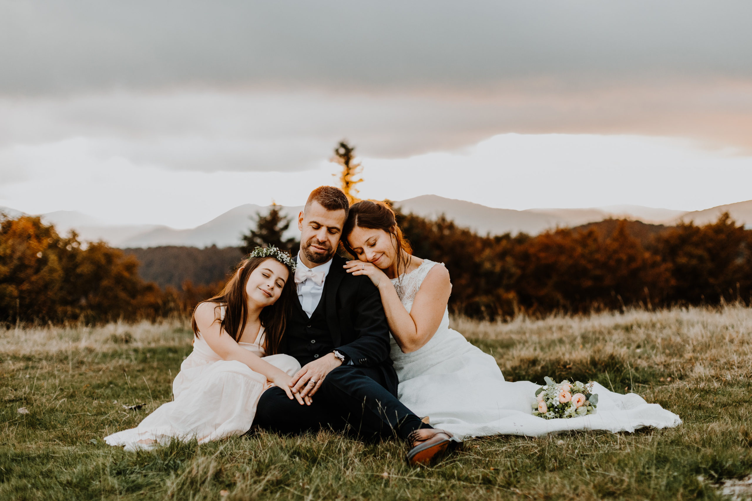 couple de mariés et leur fille assis dans l'herbe en montagne