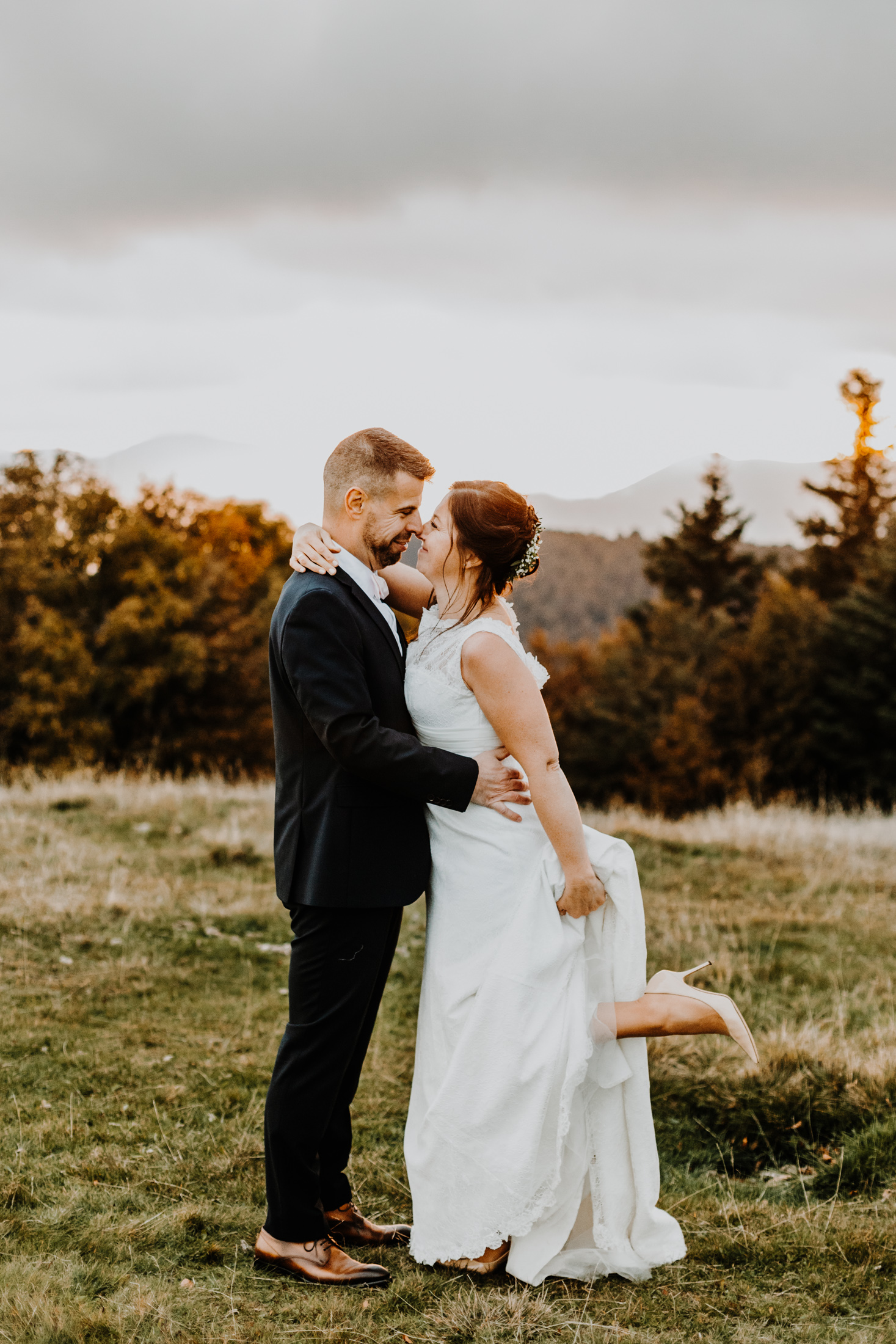 couple de mariés enlacés face à face en montagne au coucher du soleil, la mariée lèvre la jambe et la chaussure apparaît