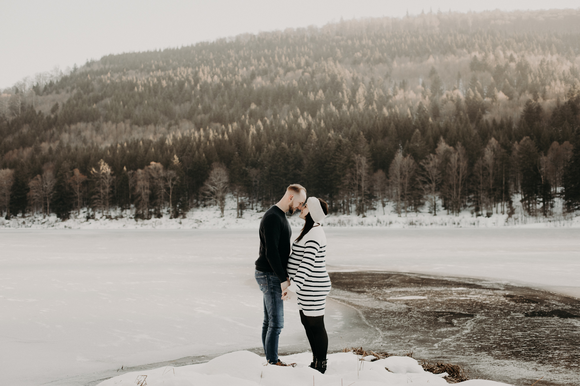 un couple de futurs parents enlacés devant un lac gelé en hiver